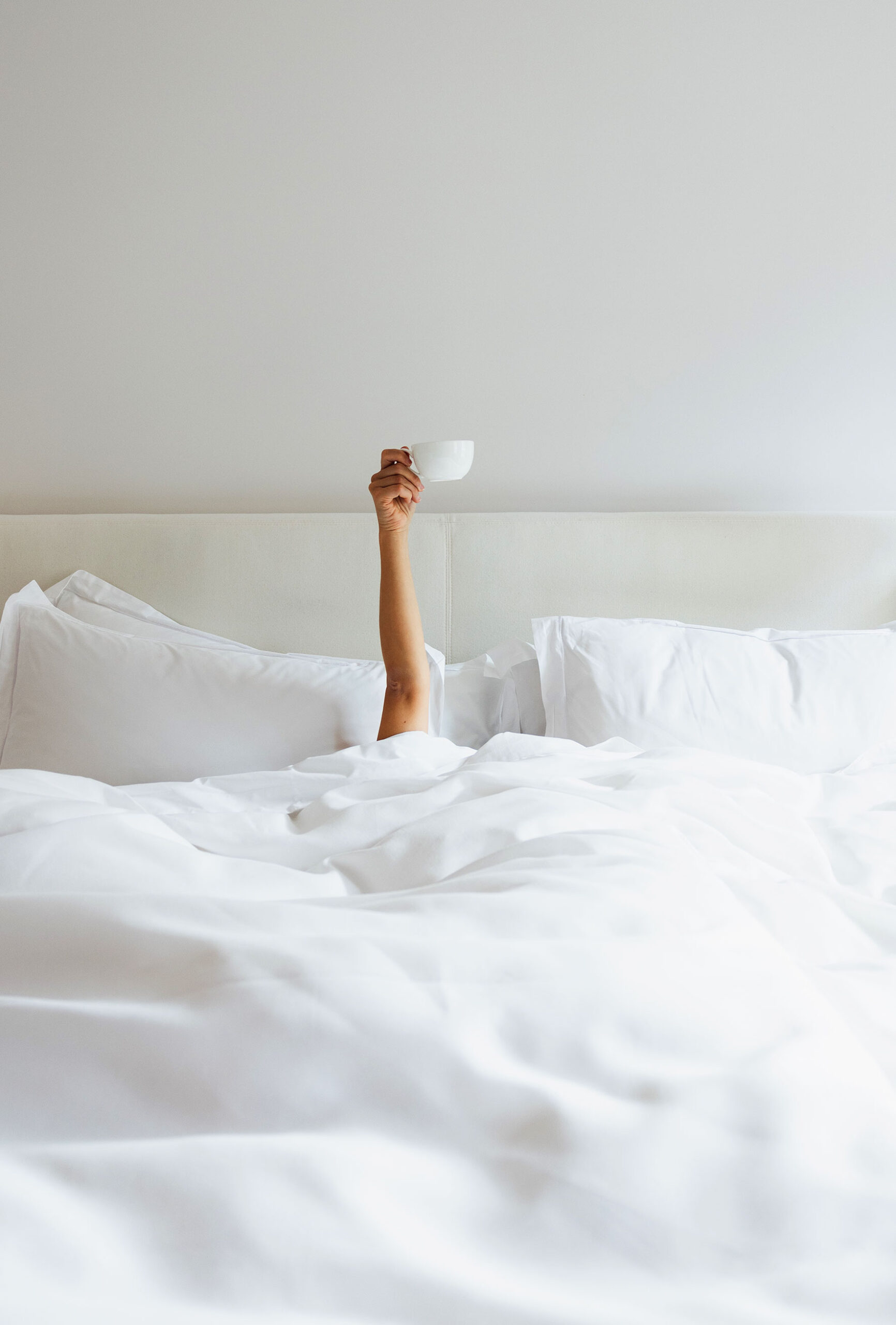 A hotel guest under the covers holding up their cup of coffee